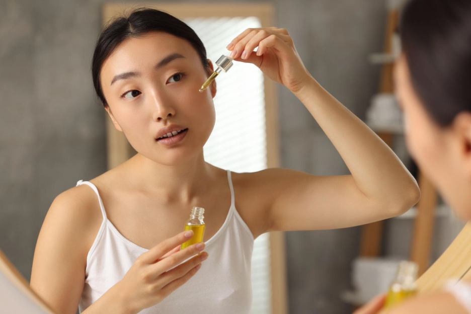 Woman applying a facial serum with a dropper in front of a bathroom mirror