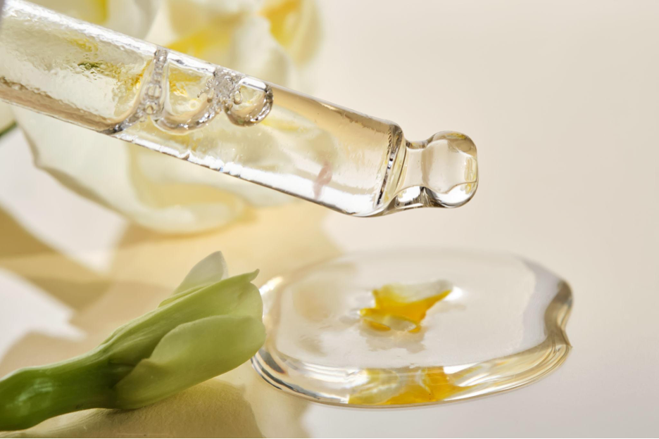 Dropper dispensing a clear botanical serum onto a glass dish beside a white flower